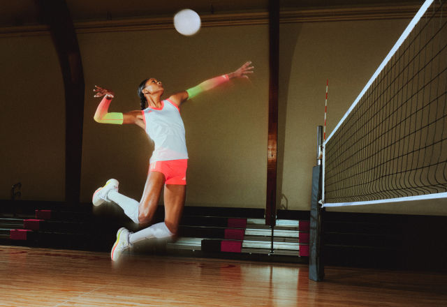 A woman in athletic wear jumps to spike a volleyball in an indoor court, with a net on the right and wooden flooring below.
