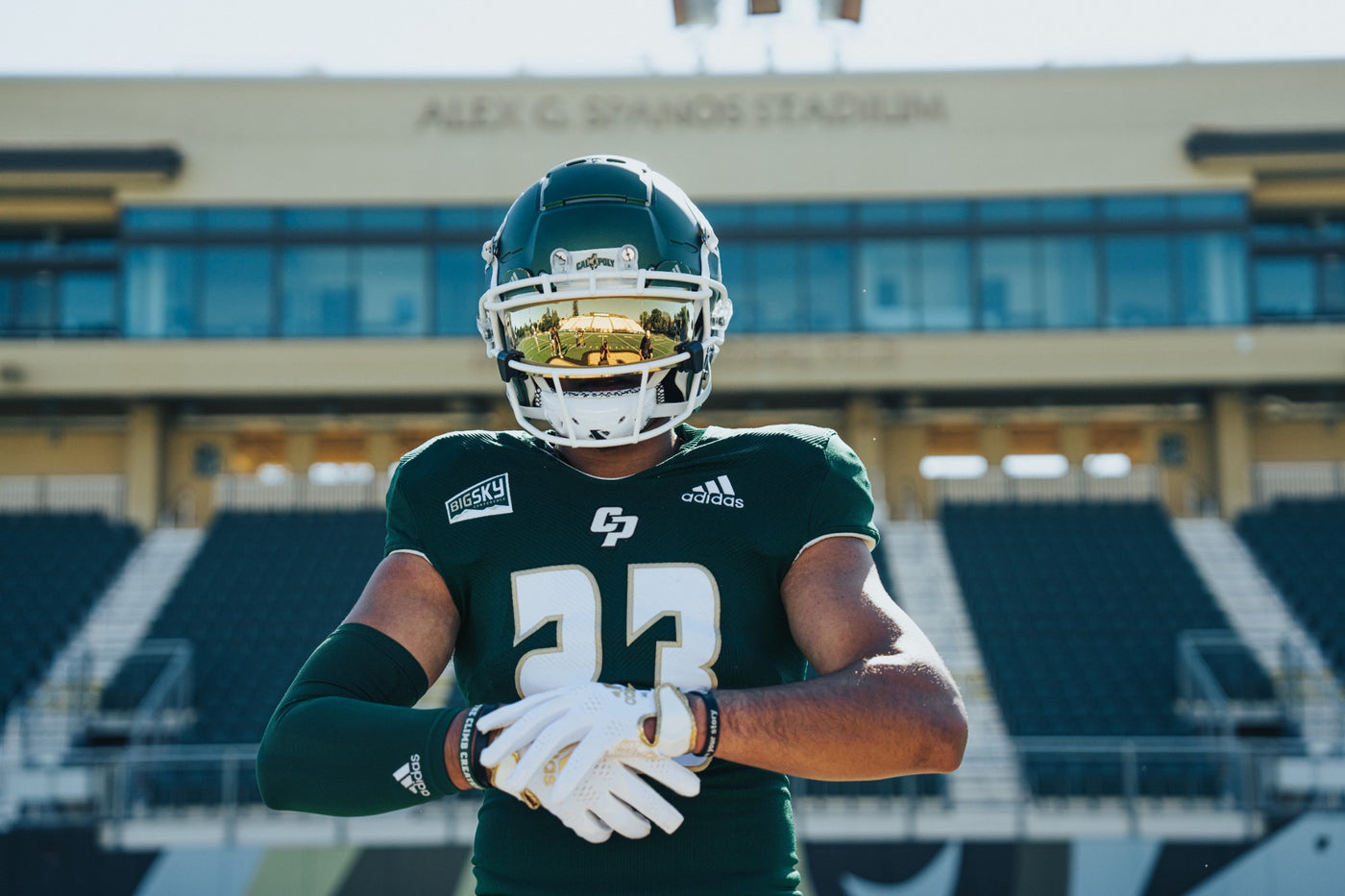 A football player in a dark green and white uniform with the number 23, wearing a reflective gold-tinted helmet and white gloves, standing in front of Alex C. Spanos Stadium.