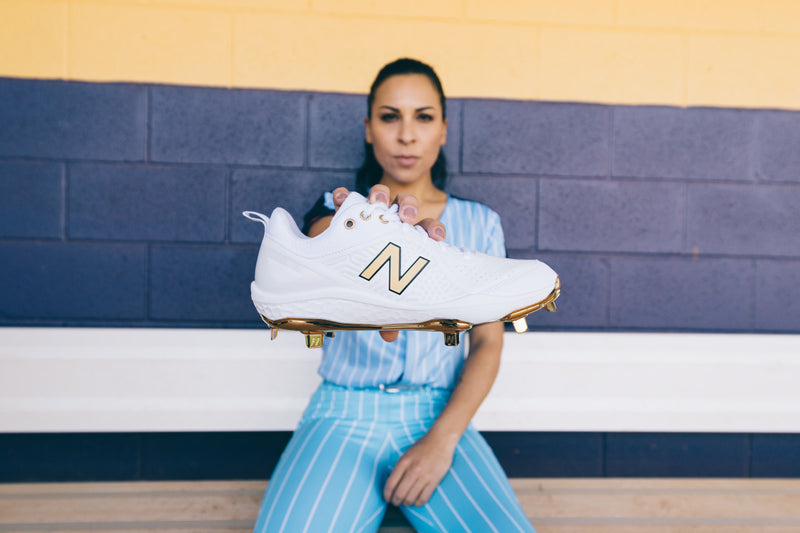 Person holding a white cleat with a gold logo against a blue and yellow wall.